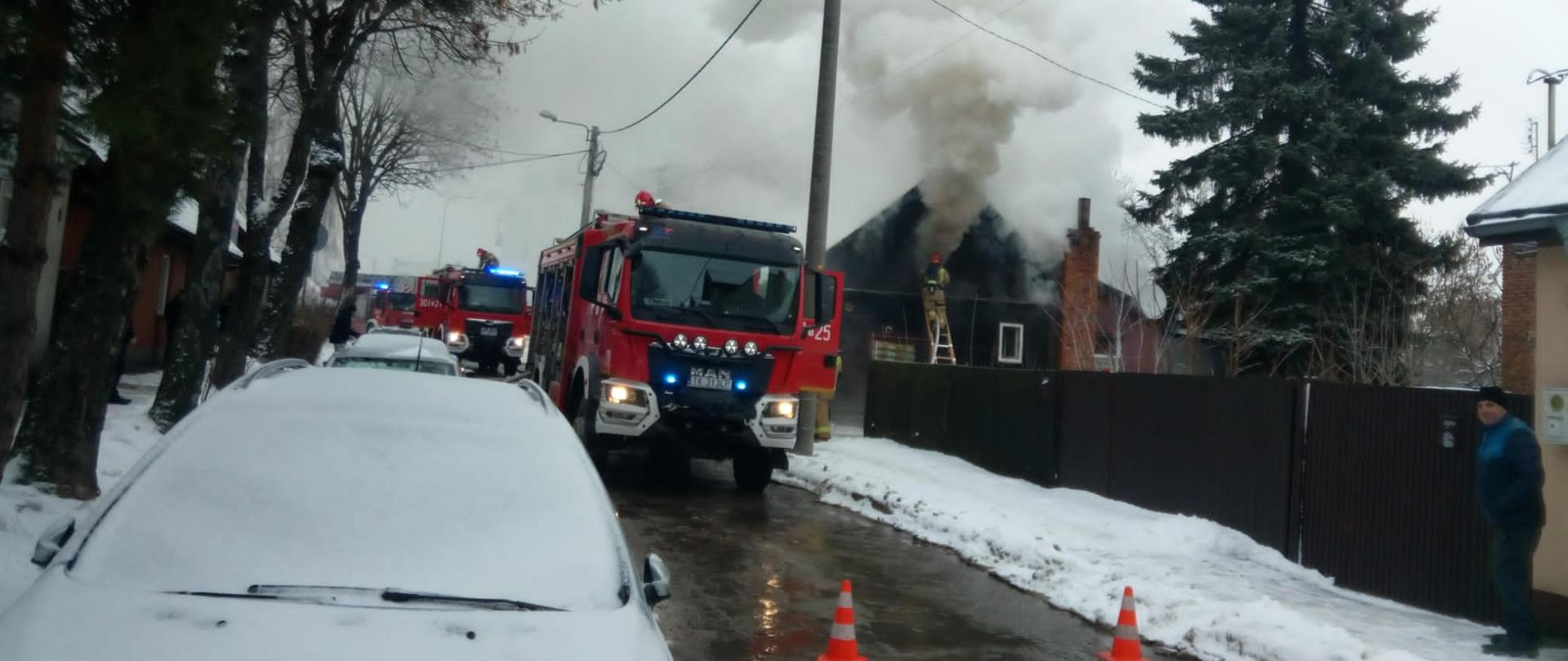 Zdjęcie przedstawia ulicę Dymińską przy której doszło do pożaru. W głębi widać budynek, z którego unoszą się kłęby dymu i pary wodnej, a przy ścianie, na drabinie stoi strażak podający wodę na poddasze. Na jezdni przy domu stoją zaparkowane trzy wozy pożarnicze a droga jest zamknięta pachołkami ostrzegawczymi.