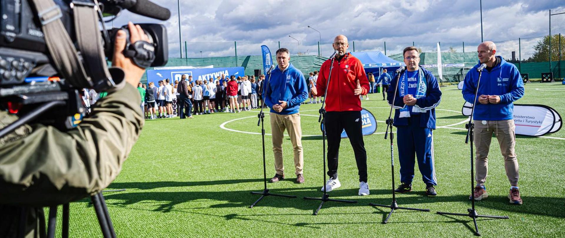 Press conference held on a football pitch. A camera can be seen on the left side of the picture. Participants stand behind microphones. From the left: Sławomir Szmal, a former top handball player and the current president of the Polish Handball Federation, Minister of Sport and Tourism Jakub Rutnicki, Michał Kmiecik, mayor of the municipality of Prażmów and Szymon Marciniak, a football referee. Children in the background.