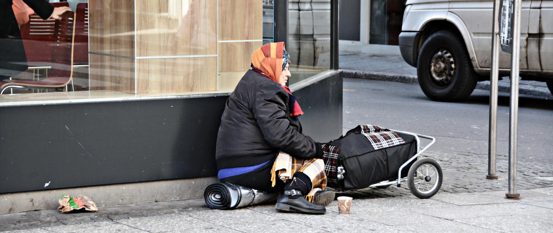 Na zdjęciu bezdomna kobieta, siedząca pod witryną restauracji. Na głowie ma kolorową chustę. Opiera się o wózek na którym ma bagaż. W tle ulica.