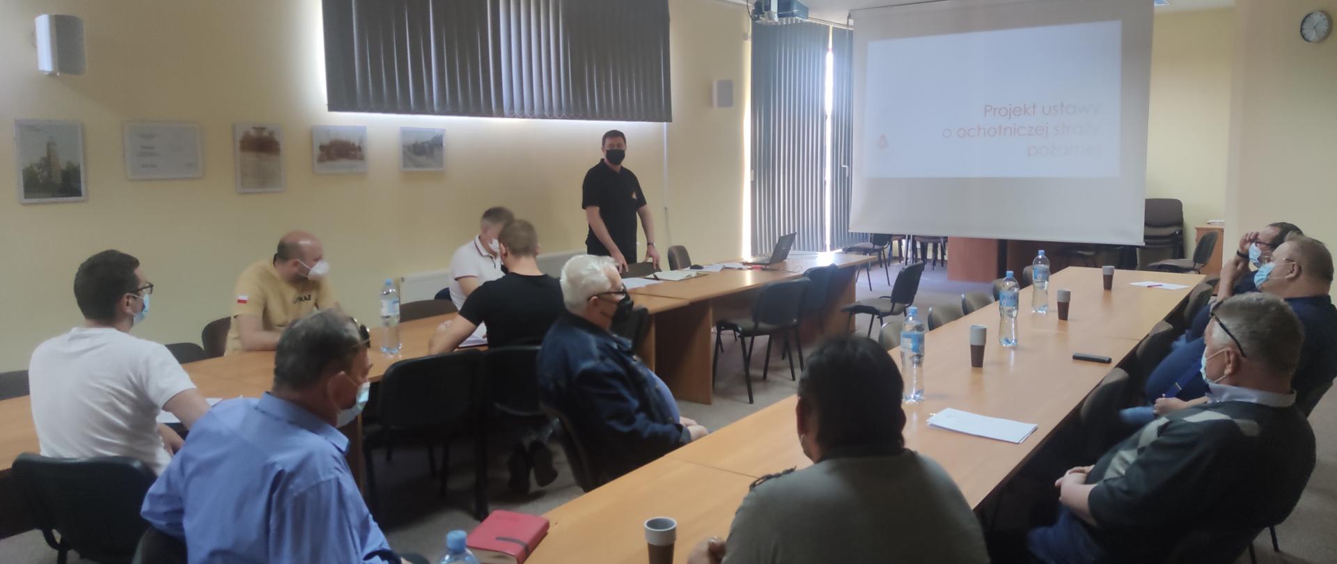 Komendant powiatowy PSP w Grodzisku Mazowiecki st. bryg. Krzysztof Tryniszewski omawia zapisy projektu ustawy o ochotniczej straży pożarnej. Przy stołach siedzą przedstawiciele ochotniczych straży pożarnych z terenu powiatu grodziskiego