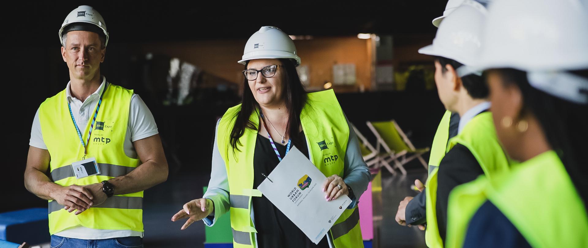 Na zdjęciu grupa ludzi stojąca w kamizelkach odblaskowych i kaskach ochronnych, w środku wiceminister Małgorzata Jarosińska-Jedynak mówiąca coś i żywo gestykulująca.