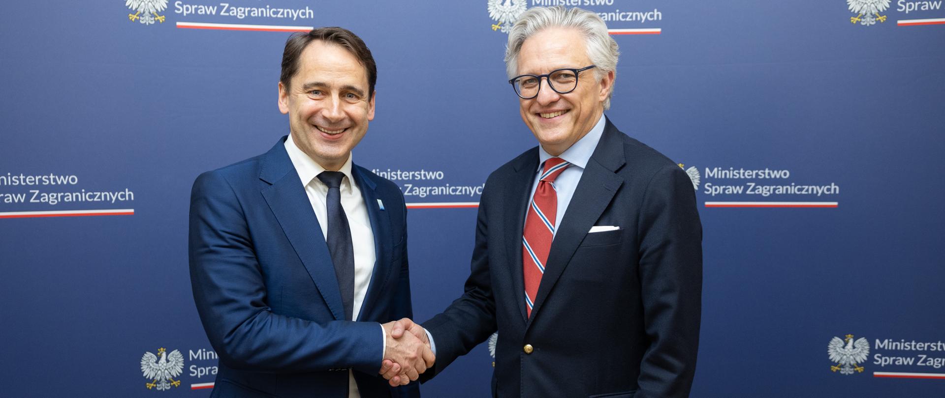  Undersecretary of State at the MFA Artur Harazim received in Warsaw Lithuania’s Vice-Minister of Foreign Affairs Sigitas Mitkus shaking hands on navy blue background with the Polish MFA logo