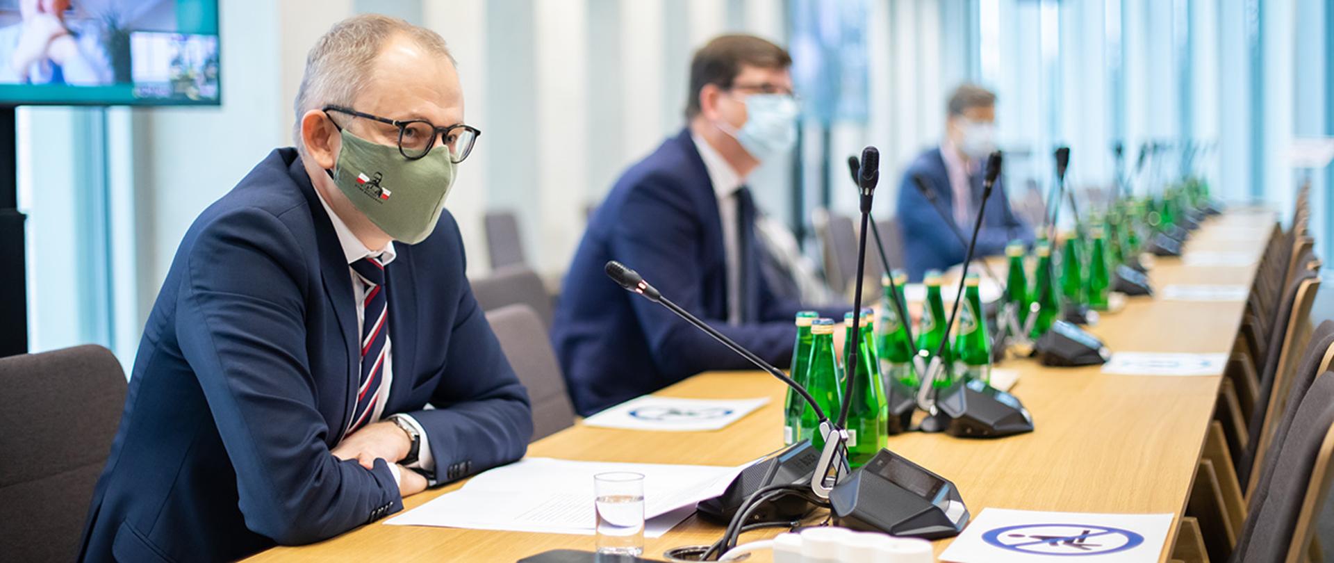 Na zdjęciu widać wiceministra Błażeja Pobożego siedzącego za stołem w maseczce zasłaniającej usta i nos. Wiceminister zabiera głos. W tle widać innych uczestników posiedzenia komisji.