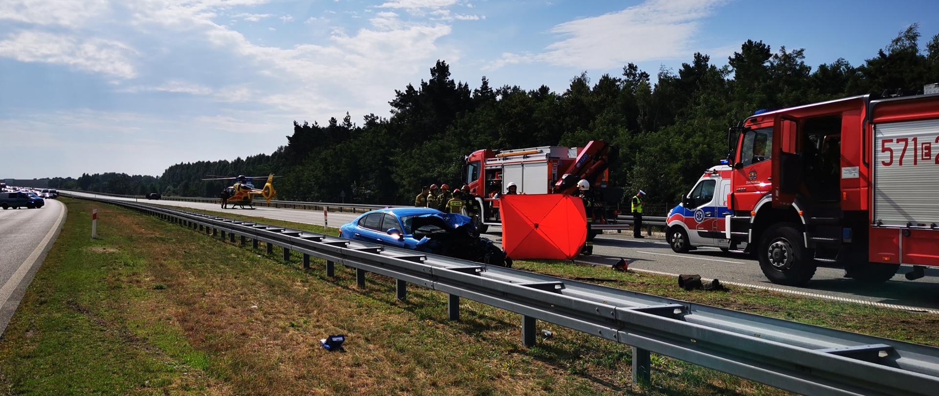 Na zdjęciu widać wozy strażackie, karetkę oraz śmigłowiec podczas zdarzenia na trasie S8, przy barierach energochłonnych widać rozbity niebieski samochód osobowy. Obok samochody strażacy trzymają czerwony parawan.