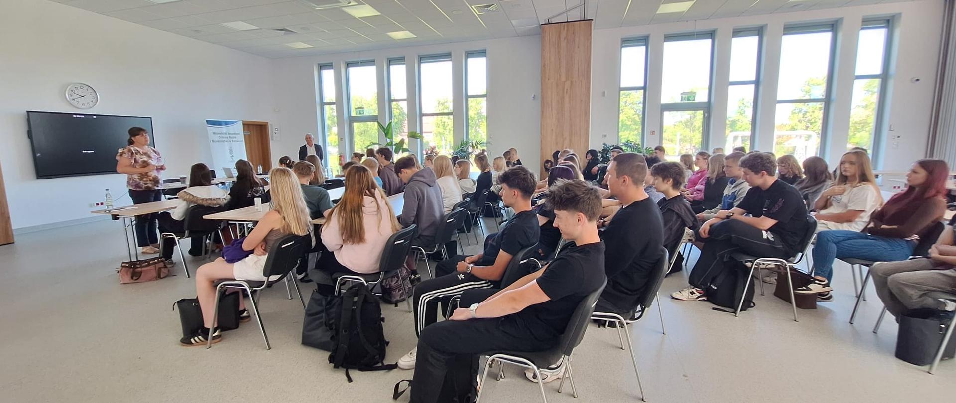 Sala lekcyjna z grupą uczniów siedzących przy stołach i słuchających prelegenta stojącego przy ekranie z kolorowym diagramem; w tle duże okna i baner instytucji.