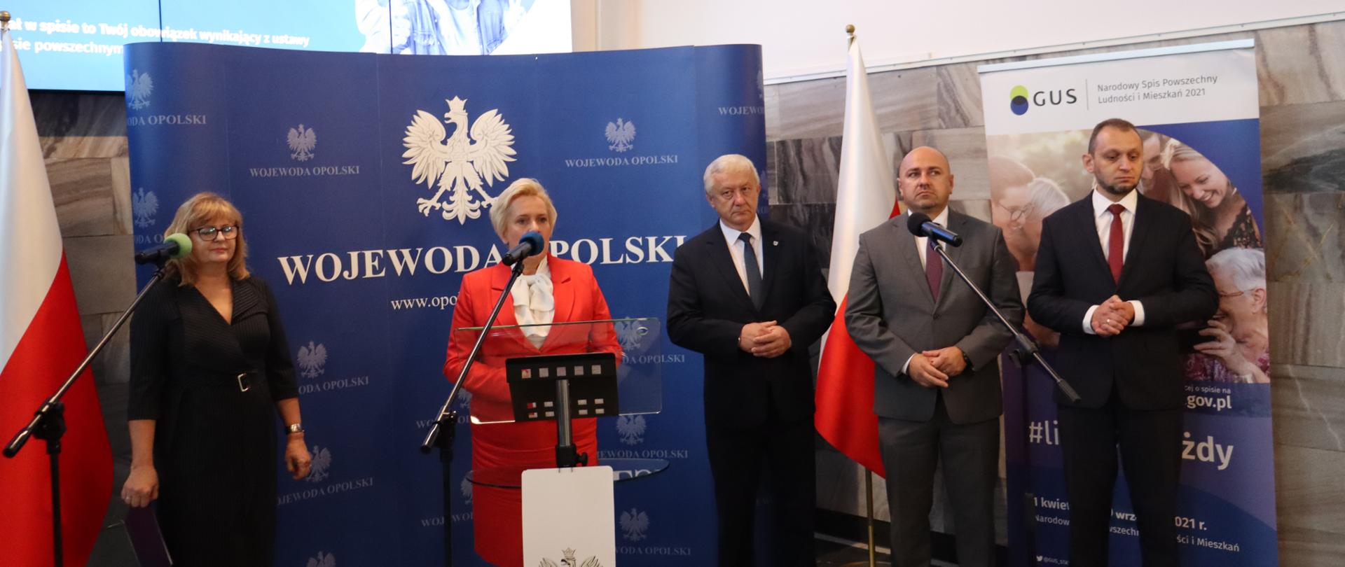 Na tle ścianki z logo wojewody opolskiego i flagami narodowymi stoją wicewojewoda opolski Teresa Barańska, dyrektor Opolskiego Urzędu Statystycznego Janina Kuźmicka oraz trzech opolskich samorządowców. Po lewej stronie kadru widać biało-czerwoną flagę. Wicewojewoda przemawia zza mikrofonu. W tle, u góry, widać fragment monitora z wyświetloną infografiką. 