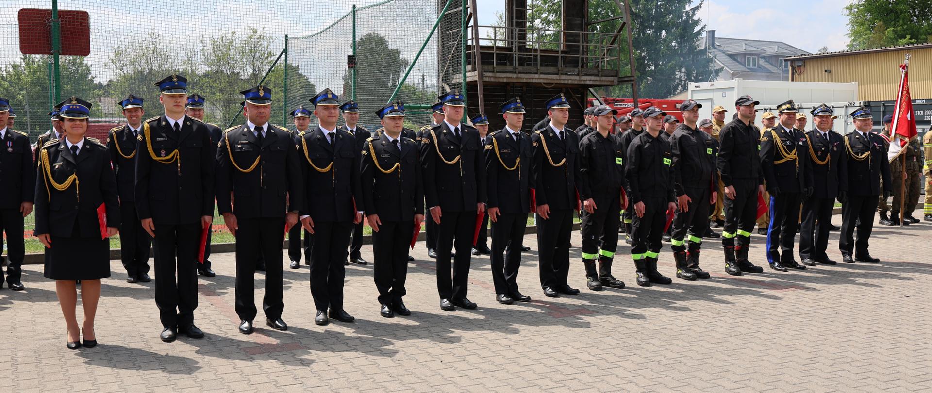Funkcjonariusze PSP oraz strażacy w ubraniach specjalnych ustawieni w szeregu podczas oficjalnych obchodów.