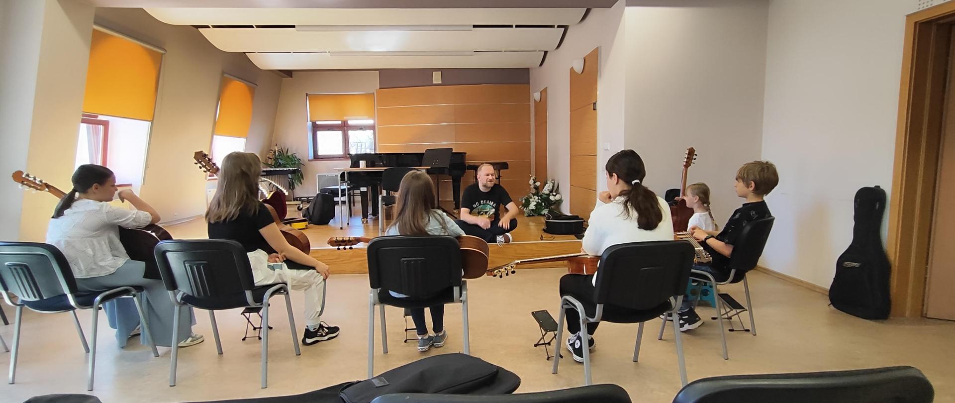 Zdjęcie na sali kameralnej. Na pierwszym planie puste krzesła na których położone są futerały. Uczniowie siedzą w półokręgu ze swoimi gitarami na kolanach. Prowadzący warsztaty siedzi po turecku na scenie sali kameralnej. Za nim fortepian, trzy okna przysłonięte roletami, w roku sali palma doniczkowa.