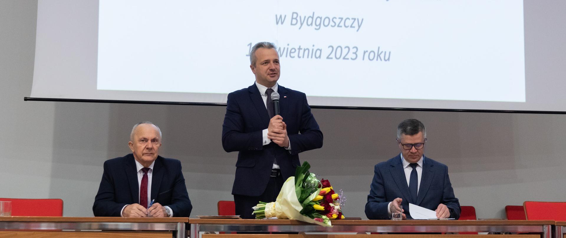 Jeden mężczyzna stojący z mikrofonem i wypowiadający się. Obok siedzi dwóch mężczyzn