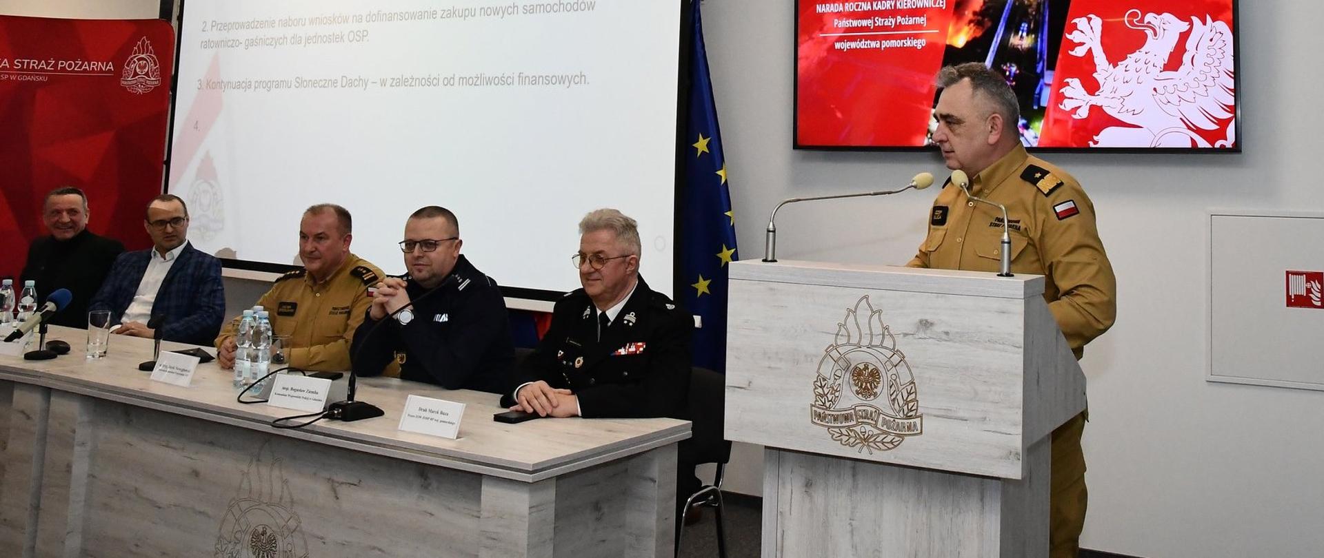 strażak w mundurze przemawia za pulpitem , kilku mężczyzn siedzi za stołem i słucha