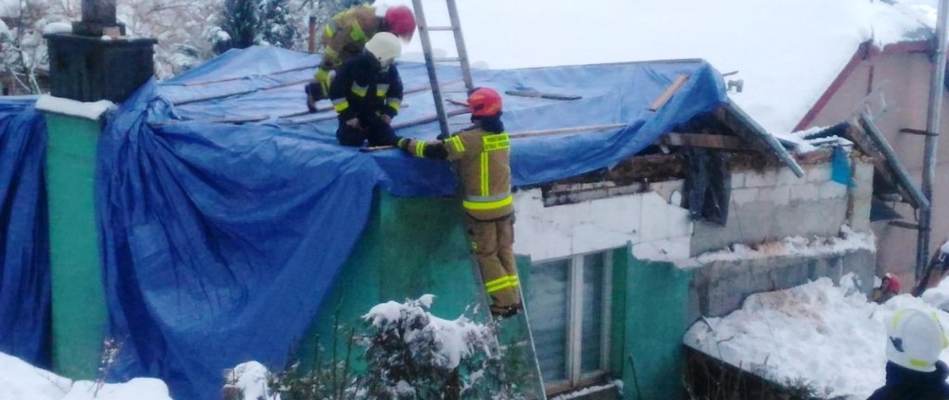 Kolorowa fotografia przedstawia na pierwszym planie część budynku z zawalonym dachem, przykrytego doraźnie niebieskimi plandekami. Na dachu dwaj umundurowani strażacy zabezpieczają listwami plandeki. obok nich na drabinie opartej do ściany budynku stoi i pomaga trzeci strażak. W tle widać inne mocno zaśnieżone budynki i innych strażaków w akcji.