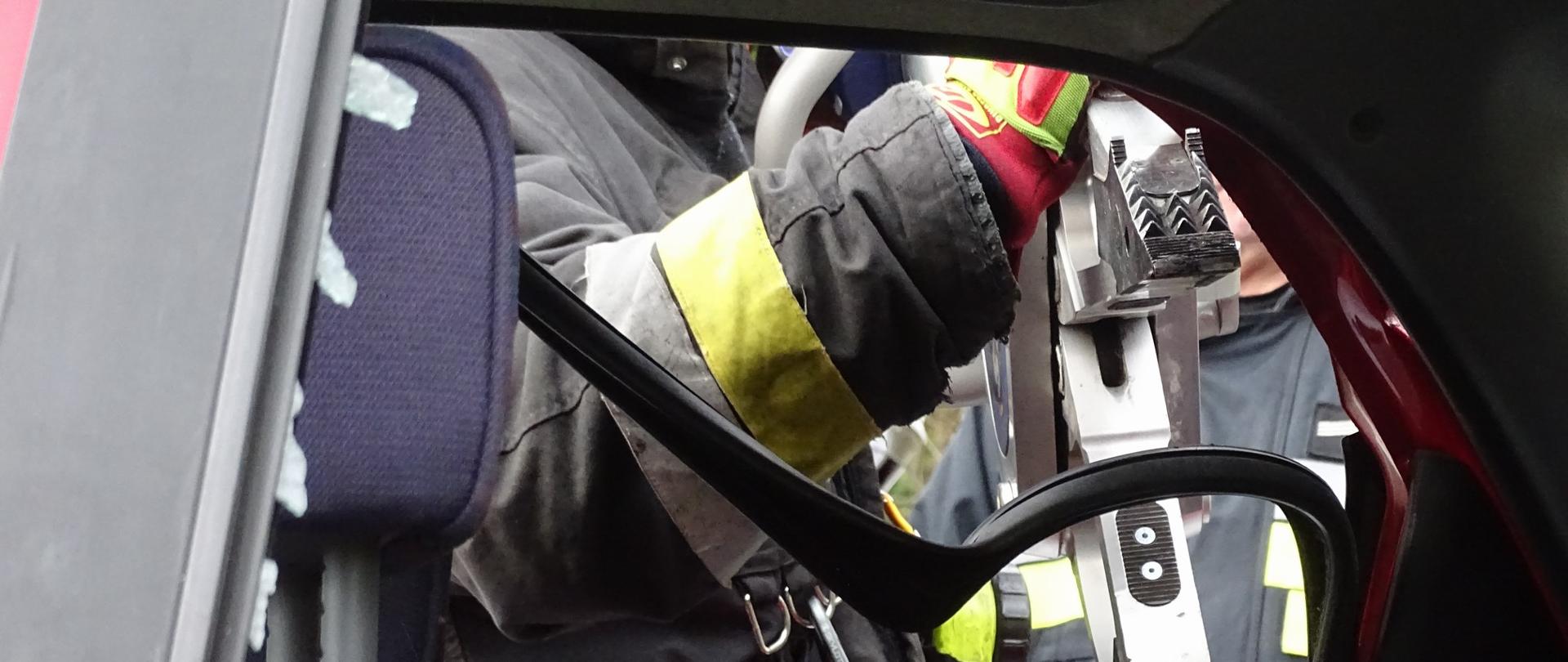 Na zdjęciu widać sylwetkę strażaka operującego rozpieraczem ramieniowym w celu otwarcia drzwi pojazdu.