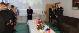 Na zdjęciu widoczni są strażacy podczas spotkania opłatkowego. Zgromadzeni stoją w świetlicy JRG wokoło stołu nakrytego białym obrusem. Na stole widoczne są potrawy wigilijne oraz napoje. W tle widać transmisję połączenia internetowego z Posterunkiem Tłuszcz.