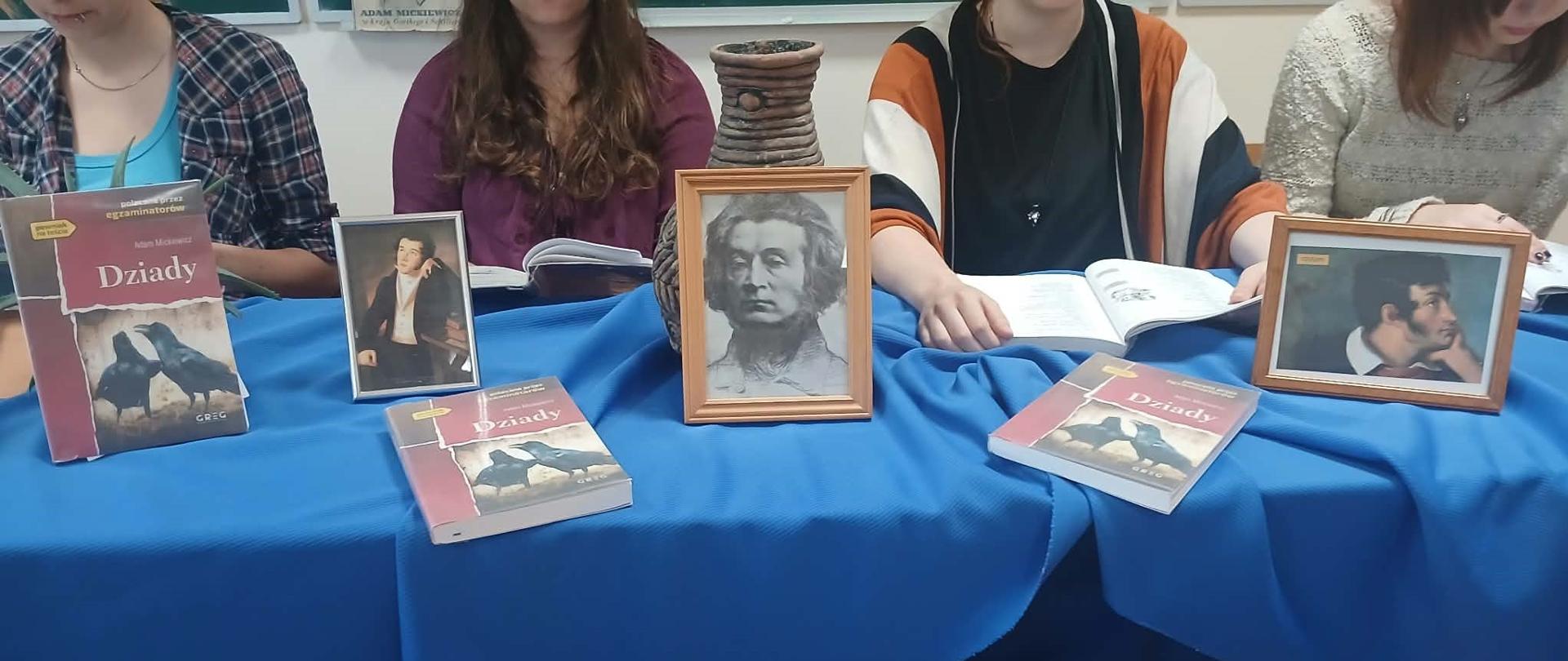 Fotografia przedstawia wystawę i wspólne czytanie Dziadów w ramach akcji Narodowe Czytanie. Centralnym motywem są książki i oprawione portrety Adama Mickiewicza wyeksponowane na stoliku, przykrytym niebieską tkaniną.