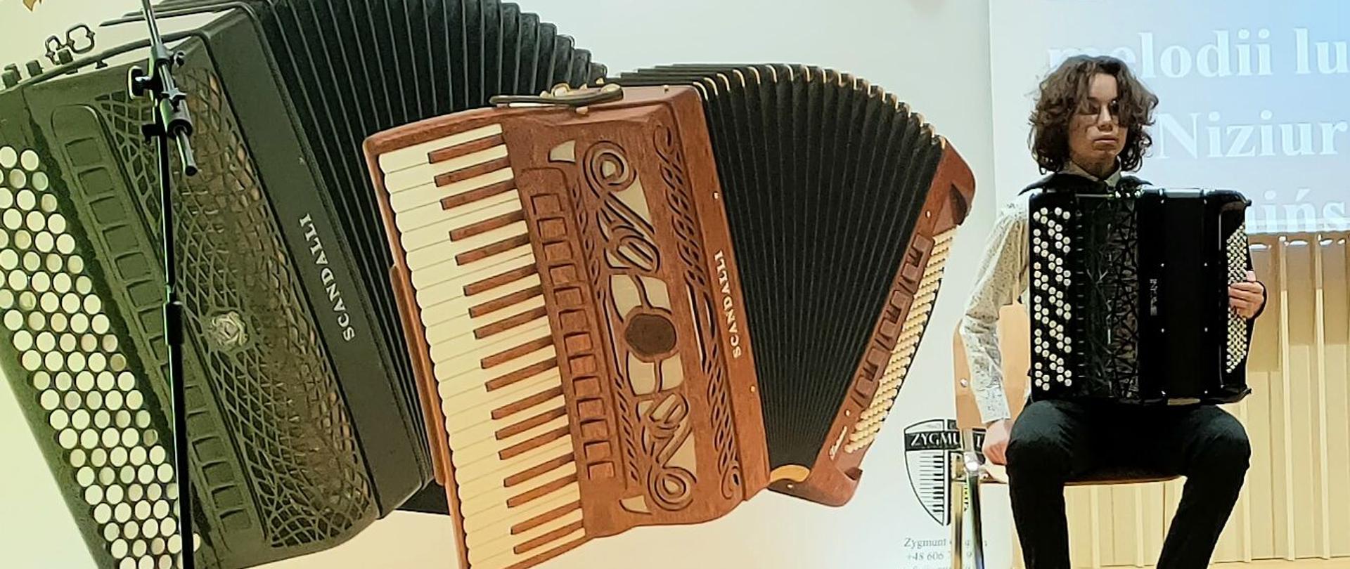 Zdjęcie kolorowe. Akordeonista siedzący na scenie. W tle dekoracja - dwa akordeony.