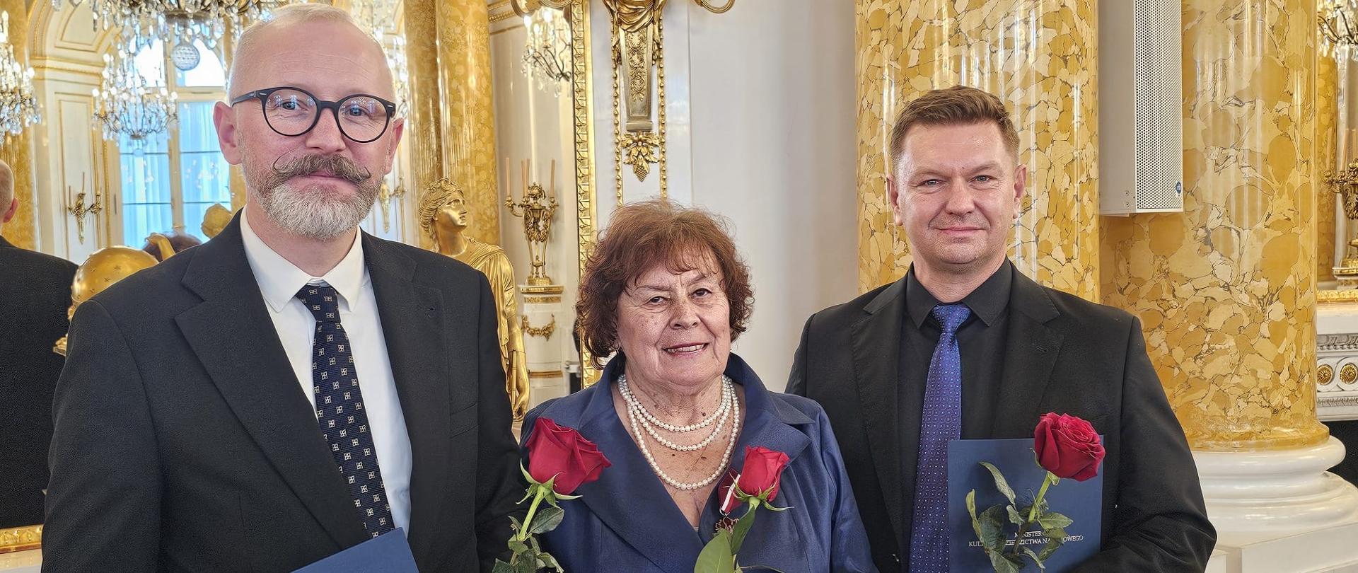Kolorowe zdjęcie na którym znajdują się trzy osoby (ich popiersia) trzymające w lewej ręce czerwoną róże oraz dwie z nich (panowie) niebieskie teczki z napisem Ministerstwo Kultury i Dziedzictwa Narodowego. Wszyscy ubrani w odświętne stroje w kolorach czerni, bieli i granatu. Od lewej Stanisław Pielczyk, Maria Warchoł-Sobiesiak i Sebastian Wieczorek