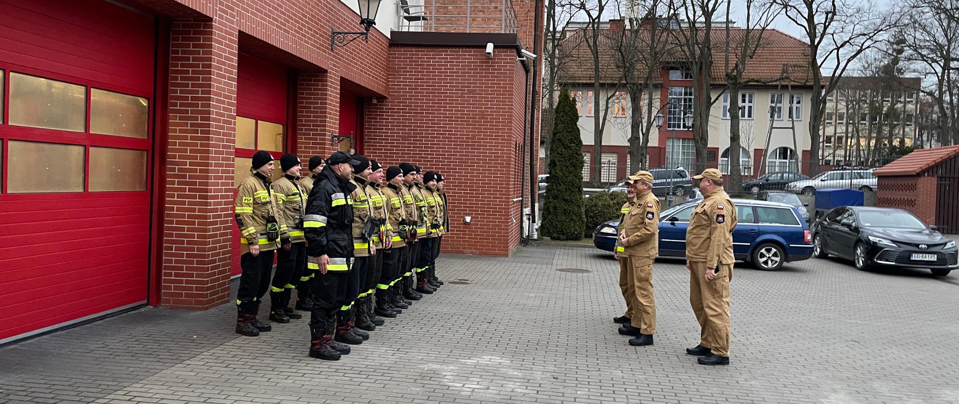 Przed garażami stoją strażacy w umundurowaniu i w dwuszeregu. Przed nimi stoi komendant wojewódzki z zastępcą i komendantem miejskim