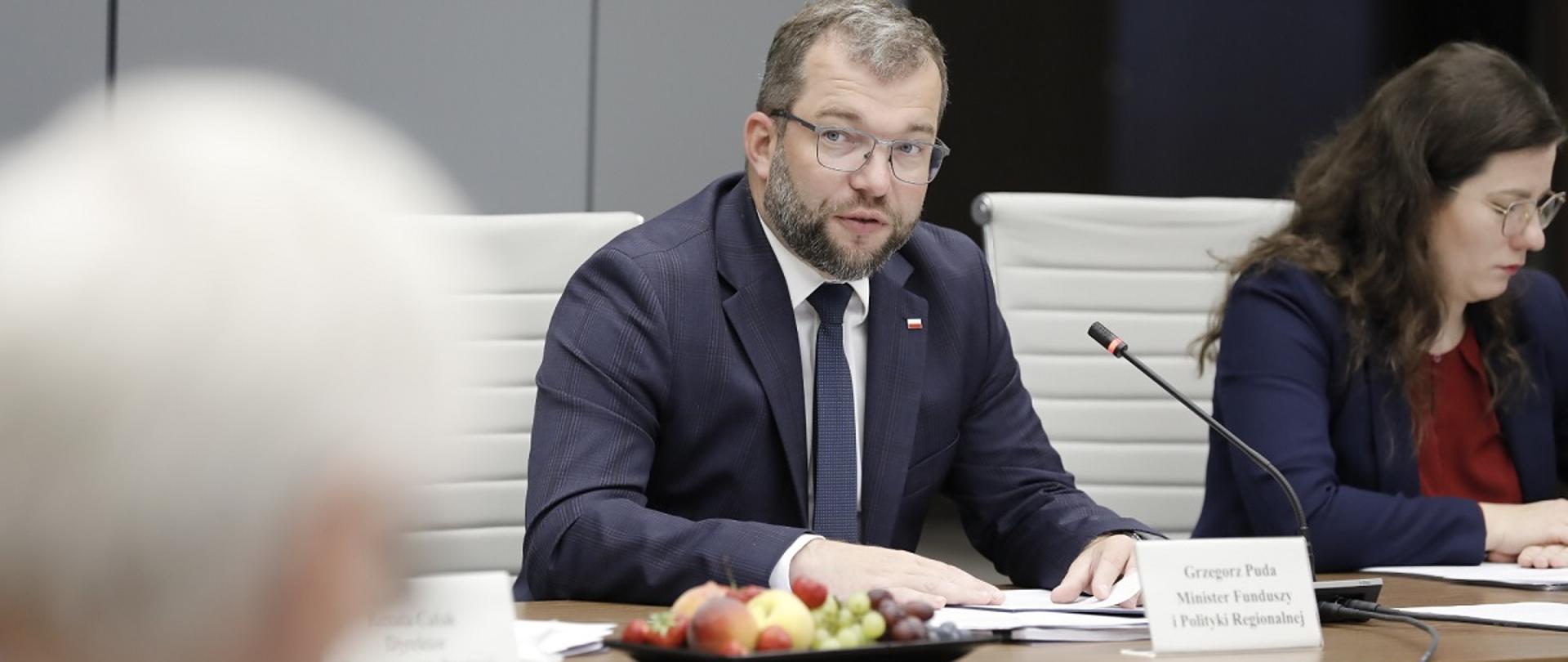 Minister Grzegorz Puda na Radzie Wykonawczej ds. wdrażania Krajowej Polityki Miejskiej 2030, minister siedzi przy stole i przemawia do mikrofony, po jego prawej stronie wicedyrektor Departamentu Strategii MFiPR Wiktoria Saganowska