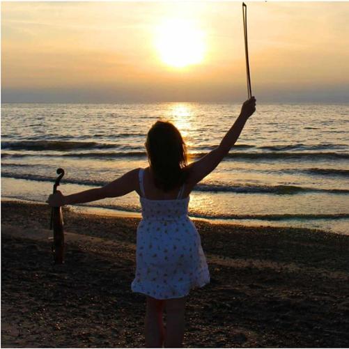Zdjęcie kolorowe. Dziewczyna w letniej białej sukience stoi trzymając skrzypce i smyczek na tle morza i zachodzącego nad nim słońca.