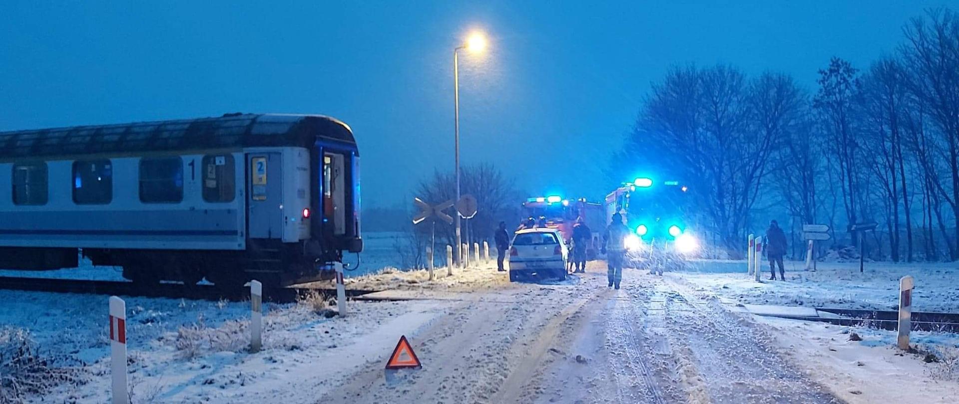 Przejazd kolejowy. Przed przejazdem trójką ostrzegawczy, na torach widoczny ostatni wagon, za przejazdem samochód który uderzył w pociąg. W tle samochody pożarnicze z włączonymi światłami alarmowymi.