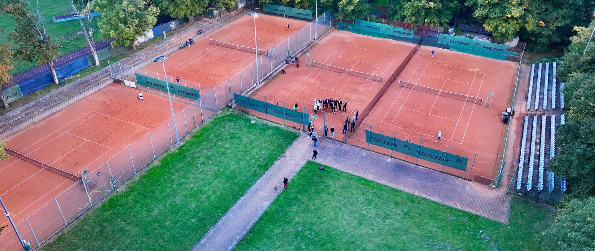 Widok z lotu ptaka na ziemne korty tenisowe. Na zdjęciu widać cztery korty i kawałek trawnika. Na jednym z kortów gromadzą się ludzie na konferencję prasową.