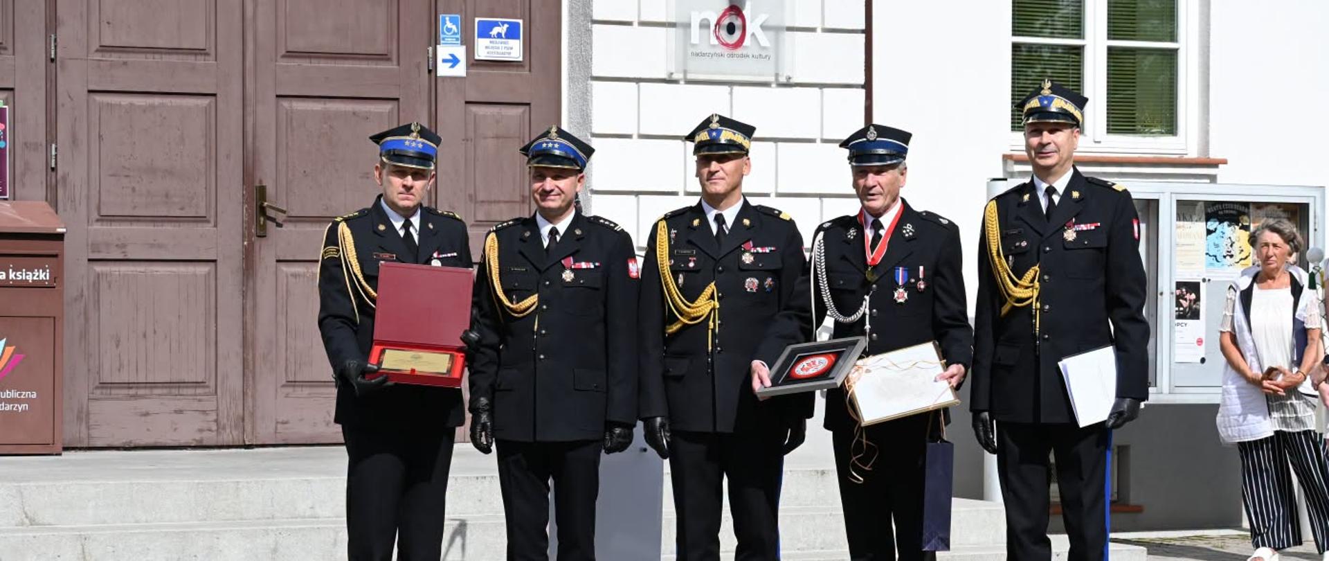 Na zdjęciu widoczna jest grupa strażaków stojących przed budynkiem podczas uroczystości. Funkcjonariusze ubrani są w wyjściowe mundury PSP z czapkami, białymi rękawiczkami i galowymi sznurami. Kilku z nich trzyma okolicznościowe dyplomy i wyróżnienia. W tle znajduje się wejście do budynku oraz pozostali uczestnicy wydarzenia. Zdjęcie oddaje podniosły charakter ceremonii oraz dumę z otrzymanych odznaczeń.