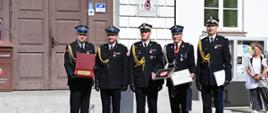 Na zdjęciu widoczna jest grupa strażaków stojących przed budynkiem podczas uroczystości. Funkcjonariusze ubrani są w wyjściowe mundury PSP z czapkami, białymi rękawiczkami i galowymi sznurami. Kilku z nich trzyma okolicznościowe dyplomy i wyróżnienia. W tle znajduje się wejście do budynku oraz pozostali uczestnicy wydarzenia. Zdjęcie oddaje podniosły charakter ceremonii oraz dumę z otrzymanych odznaczeń.