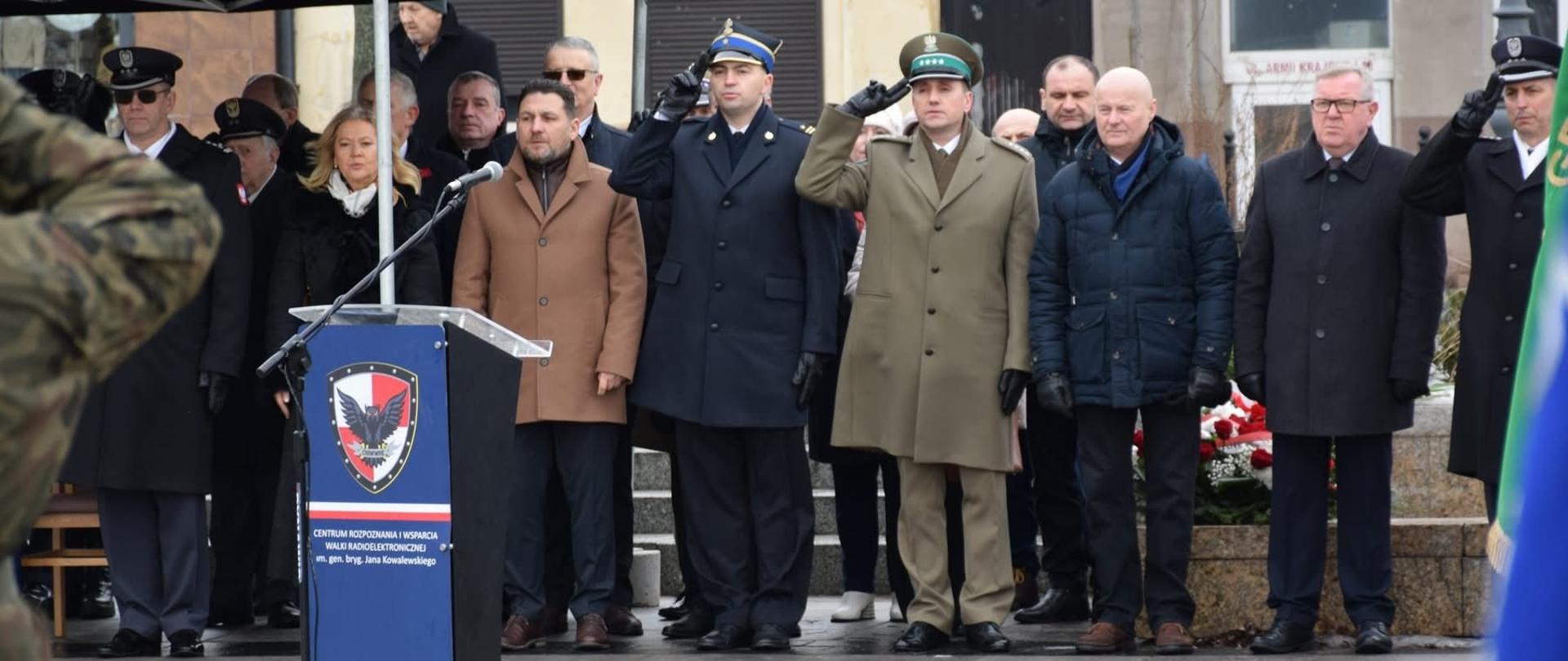 Przed mikrofonem i mównicą stoi grupa osób, w tym mężczyźni w mundurach oraz osoby w ciemnych płaszczach. Kilku z nich oddaje salut, przykładając prawą rękę do czapki. Na mównicy widoczny jest emblemat wojskowy. Za nimi stoją inni uczestnicy uroczystości. Atmosfera jest poważna i oficjalna.
Całość przedstawia oficjalne obchody lub ceremonię upamiętniającą, podczas której składane są wieńce i oddawany jest hołd.