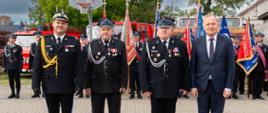 Czterej mężczyźni — trzej w mundurach strażackich oraz jeden w eleganckim garniturze — stoją w jednym rzędzie na wybrukowanym placu. Za ich plecami znajdują się strażacy trzymający sztandary oraz pojazdy pożarnicze.