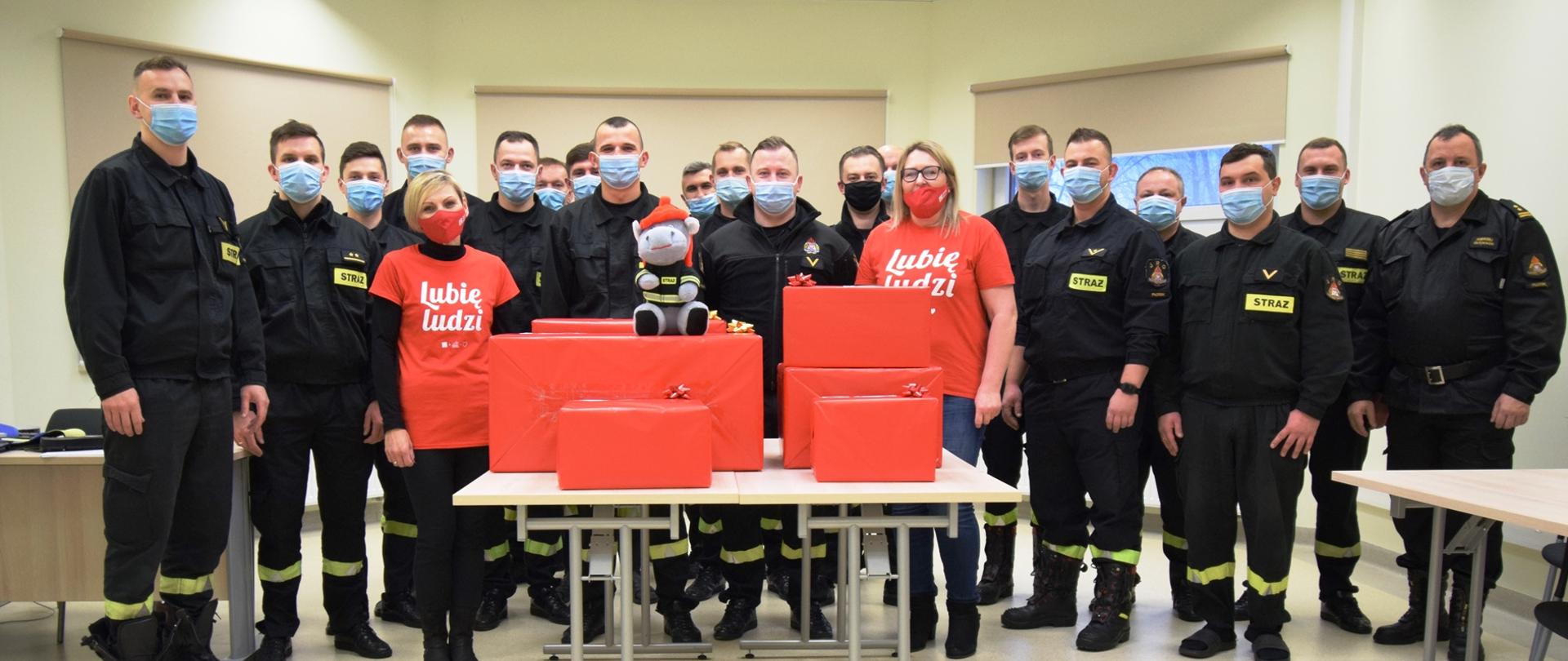 Grupa osób zaangażowana w akcję szlachetna paczka. W środku paczki i wolontariusze Szlachetnej Paczki w czerwonych koszulkach, z wokół strażacy w ubraniach koszarowych koloru czarnego.
