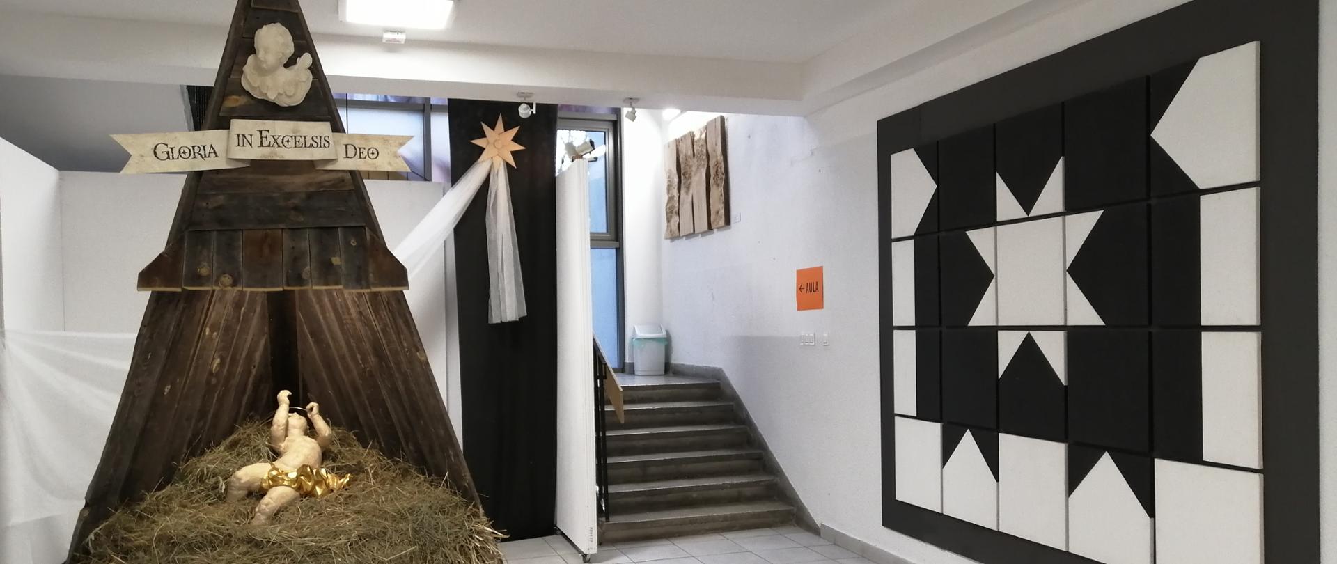 Drewniana, wysoka szopka w kształcie trójkąta zakończona rzeźbą, popiersiem anioła i napisem "Gloria in excelsis Deo". W środku szopki na sianie leży rzeźba Jezusa. Za nią wisi biała szarfa zakończona gwiazdą. Obok na ścianie wisi abstrakcyjny obraz J. Kapusty. Kwadraty zostały ułożone w ten sposób, że tworzą gwiazdę i fragment murów.