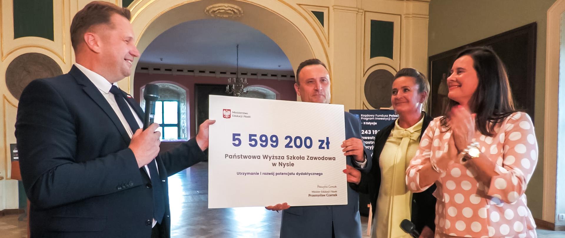 Minister stoi obok grupy osób - dwóch kolorowo ubranych kobiet i mężczyzny w garniturze, który trzyma duży symboliczny czek z napisem 5 599 200 zł.