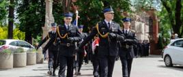 Fotografia przedstawia uroczysty przemarsz strażaków w czarnych mundurach galowych, idących w zorganizowanym szyku przez ulicę. Na pierwszym planie idą dwaj wyprężeni dowódcy, a za nimi podążają kolejni funkcjonariusze oraz poczet sztandarowy. Akcja dzieje się w słoneczny dzień w przestrzeni miejskiej, w tle widać kościół oraz drzewa.