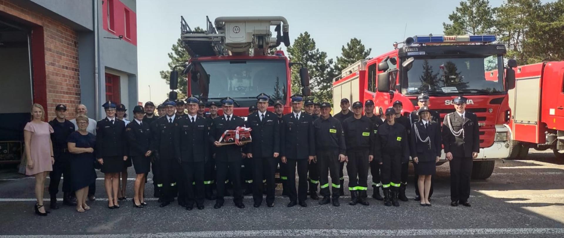 Na zdjęciu znajdują się strażacy ustawieni przed samochodami pożarniczymi na tle budynku komendy