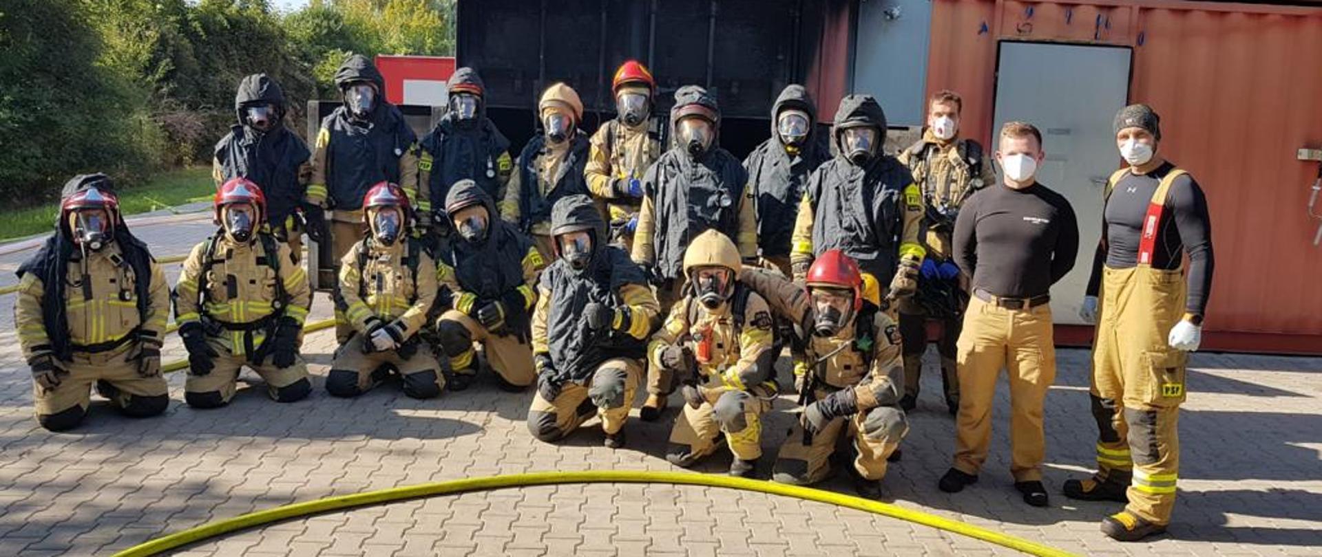 Zdjęcie przedstawia uczestników warsztatów szkoleniowych instruktorów gaszenia pożarów wewnętrznych. Strażacy w mundurach we wspólnym zdjęciu podczas ćwiczeń przed komorą rozgorzenia.