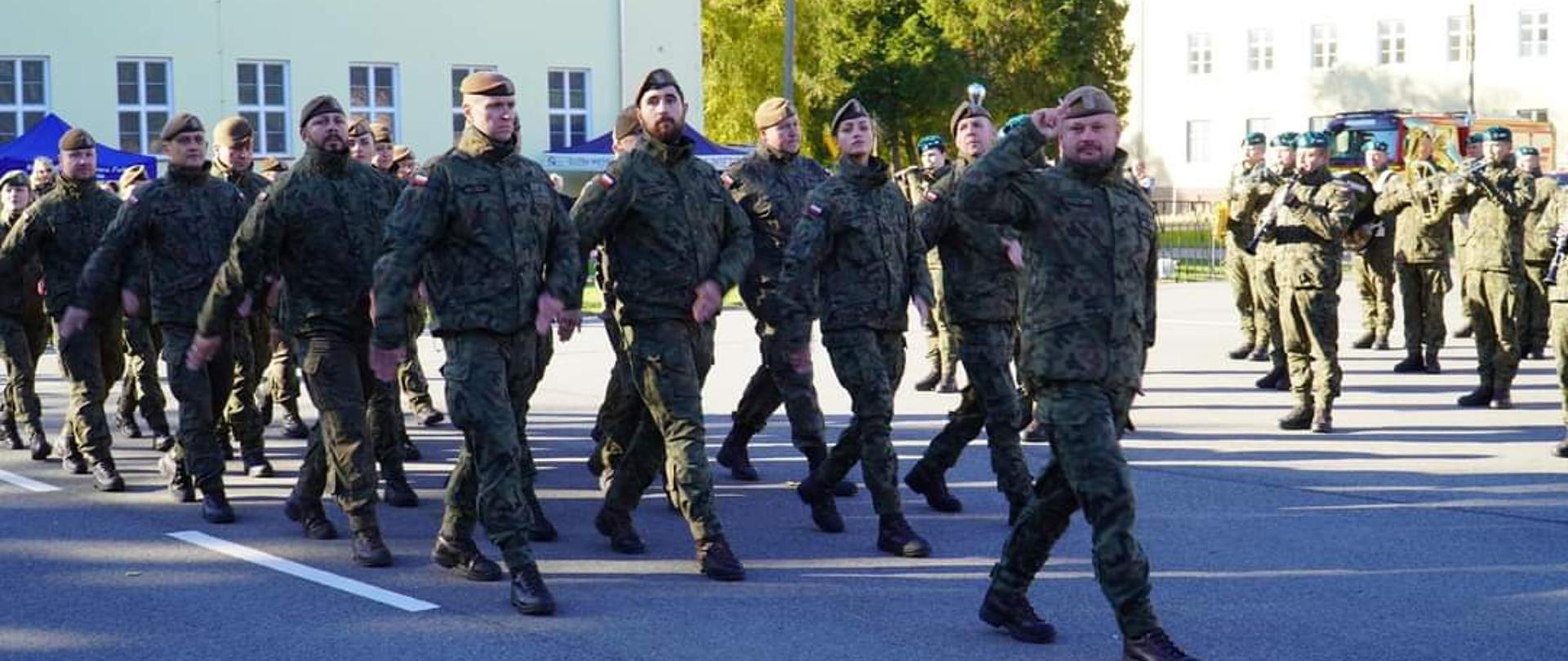 Maszerują żołnierze. Słonecznie. W tle budynki.