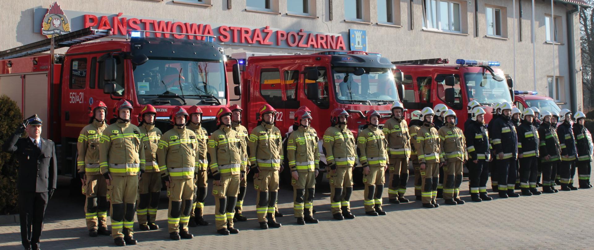 strażacy stoją w dwuszeregu przed budynkiem komendy powiatowej w pleszewie, za nimi widoczne wozy strażackie