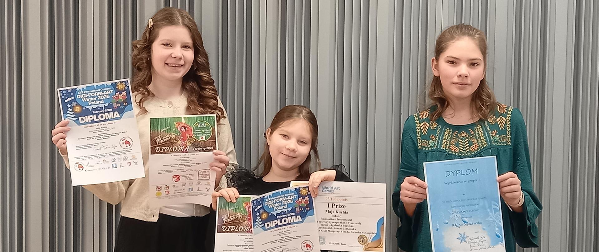 Uczennice na tle szarej kotary trzymają w rekach dyplomy