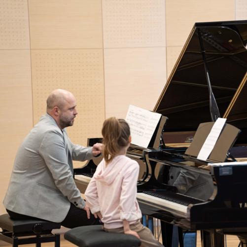 zdjęcie przedstawia prowadzącego warsztaty siedzącego przy drugim fortepianie głębiej na scenie sali koncertowej omawiającego tekst nutowy utworu na pulpicie, na pierwszym planie uczennica siedzi słuchając przy pierwszym fortepianie