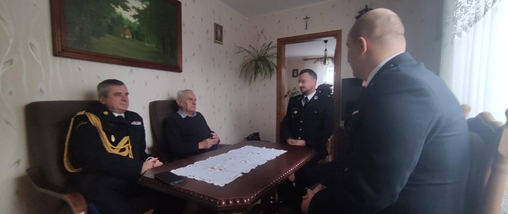 Strażak Państwowej Straży Pożarnej strażacy ochotniczej straży pożarnej oraz mężczyzna siedzą na fotelach przy ławie.