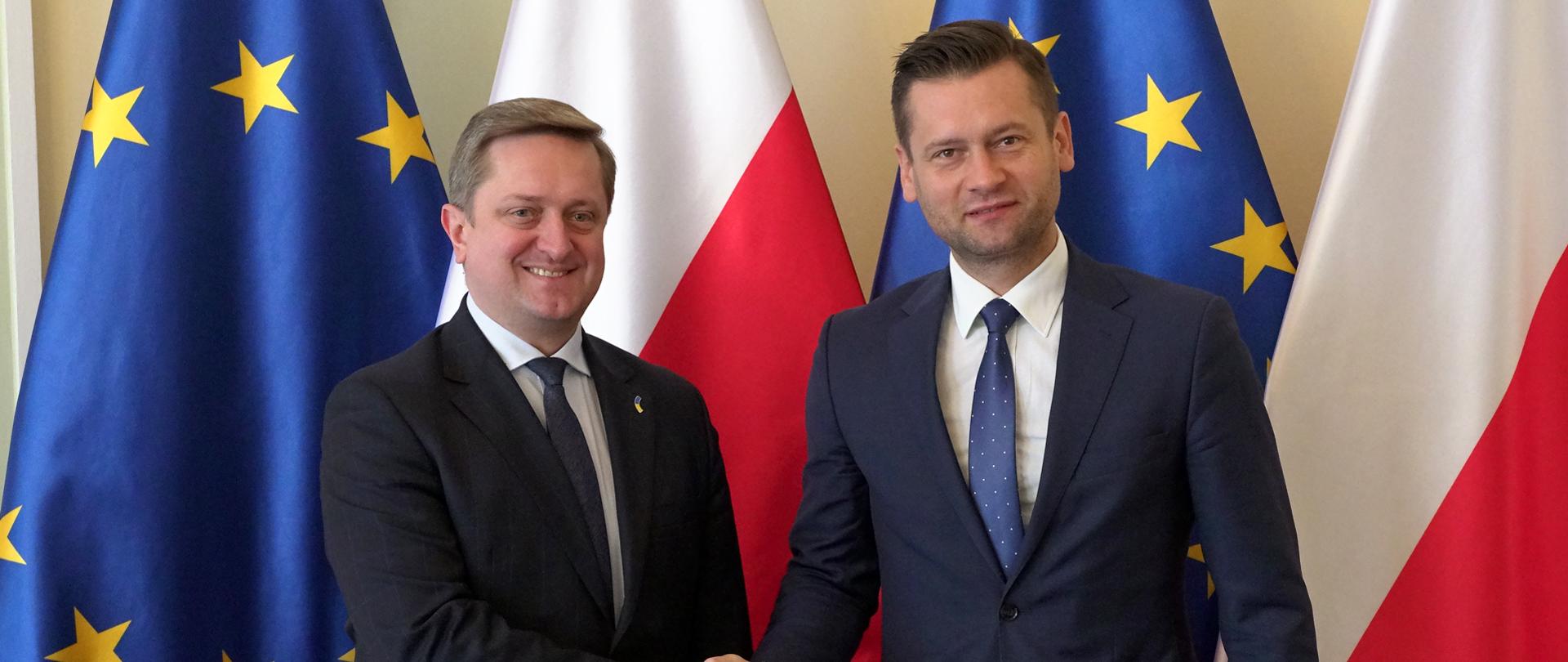 Spotkanie ministra Bortniczuka z Ambasadorem Nadzwyczajnym i Pełnomocnym Ukrainy w Polsce - minister z ambasadorem podają sobie dłonie na tle flag polski i UE