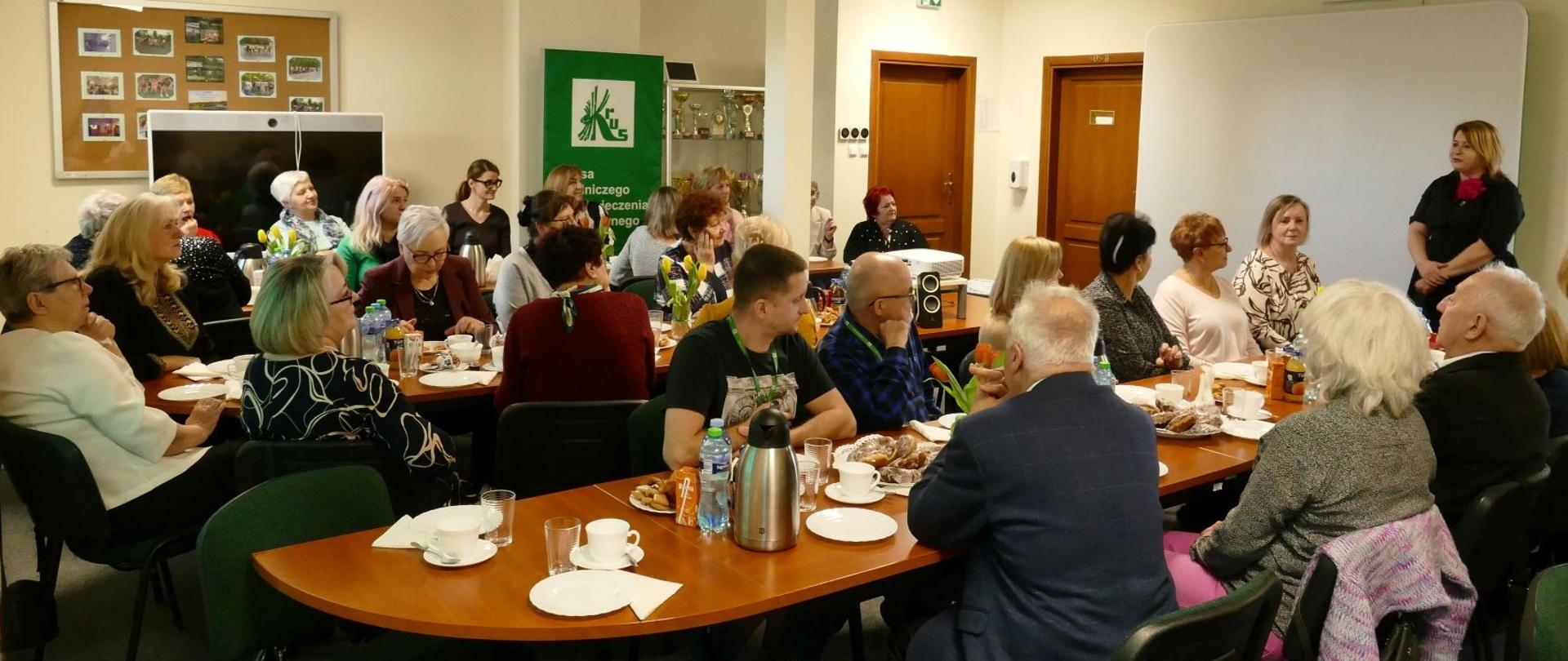 Na sali konferencyjnej przy stołach, na których stoją termosy z kawą i ciastka siedzi kilkadziesiąt osób. Po prawej stronie sali stoi kobieta, która przemawia do zebranych osób.