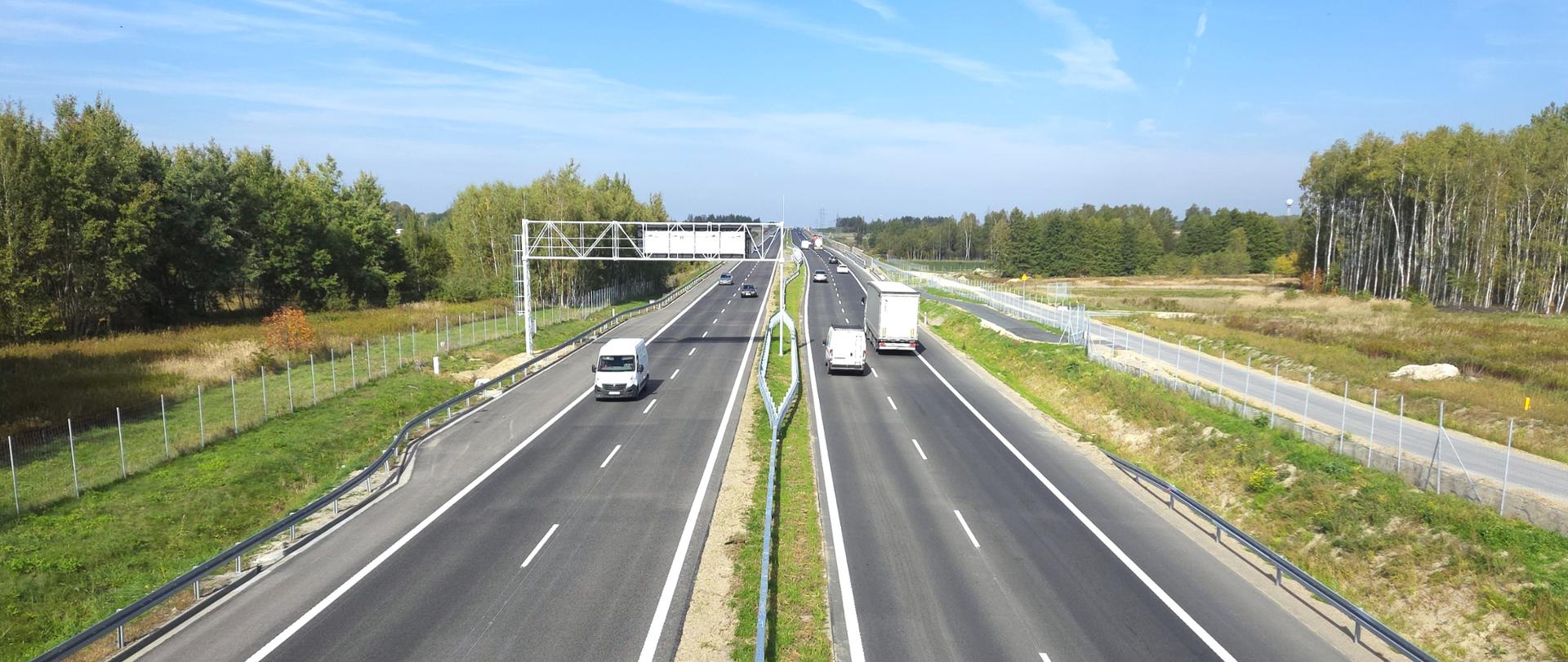 Widok z góry na szeroką, dwujezdniową drogę ekspresową biegnącą przez tereny leśne. Po obu stronach jezdni porusza się kilka samochodów osobowych i ciężarowych. Drogę otaczają zielone pobocza, ogrodzenia oraz las, a nad całością rozciąga się bezchmurne, jasnoniebieskie niebo.