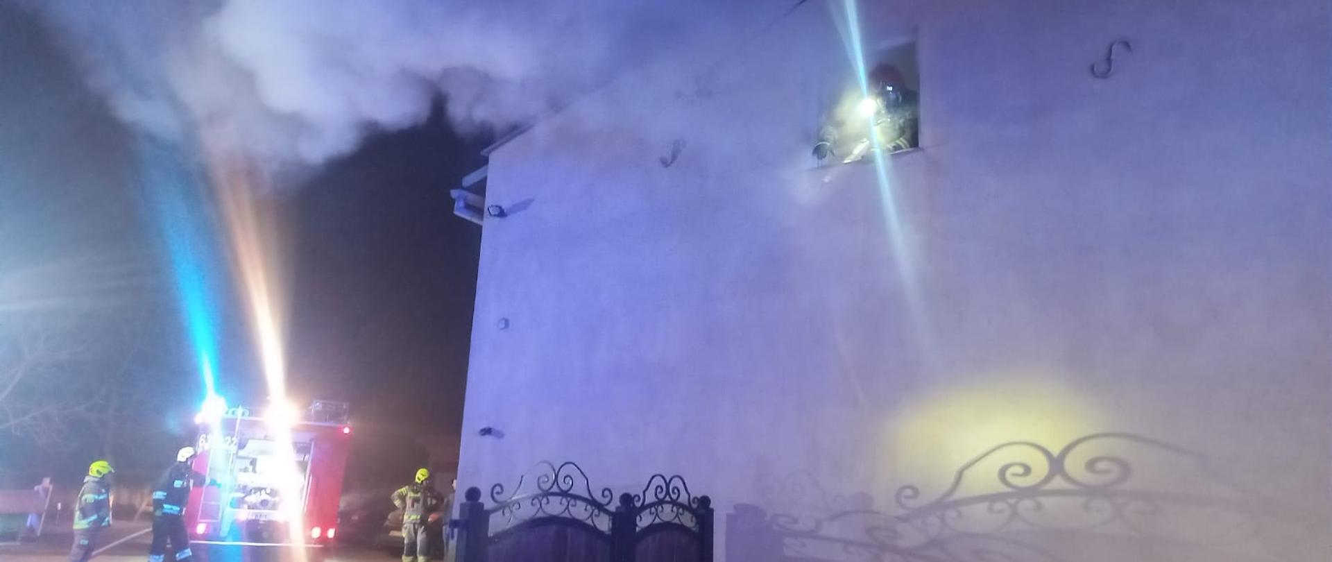 Na zdjęciu widać budynek, w którym doszło do pożaru oraz wóz strażacki i strażaków 