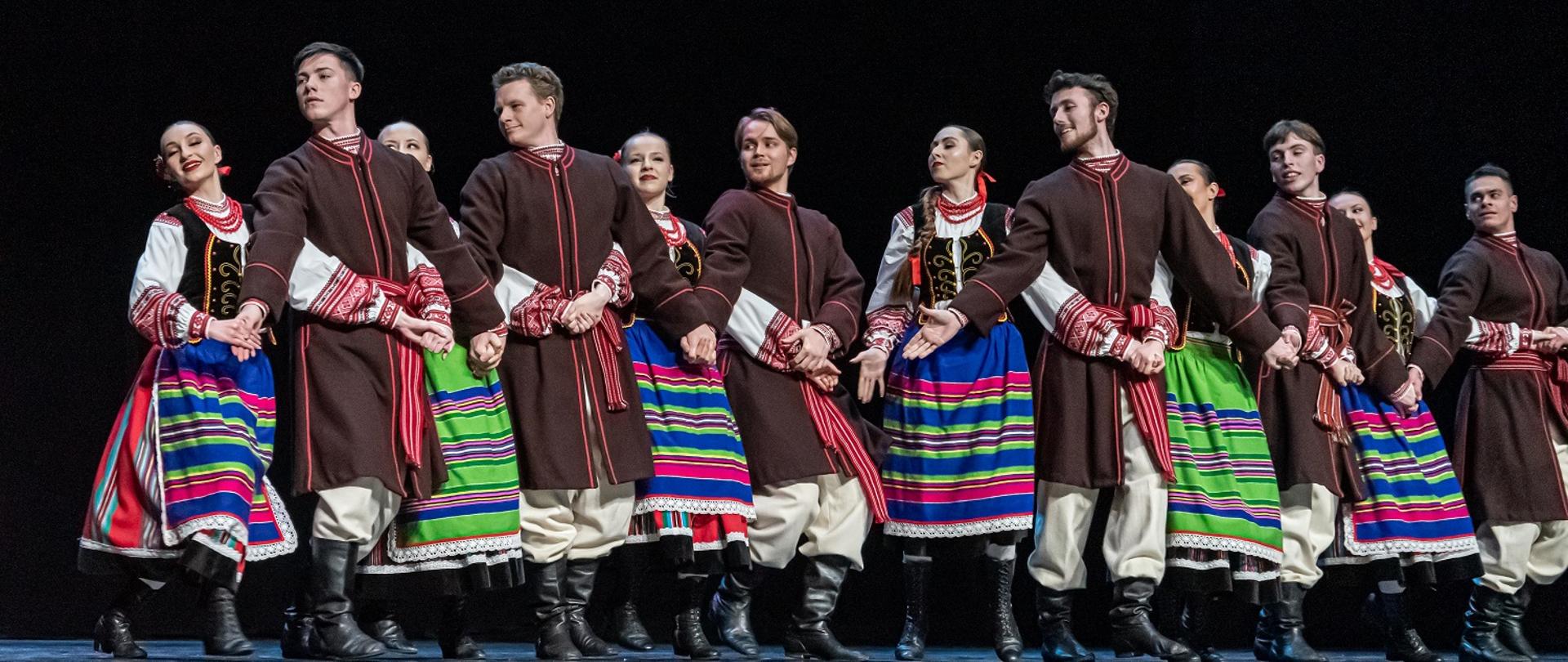 Na scenie tańczy grupa taneczna, ubrani są w regionalne stroje ludowe. 