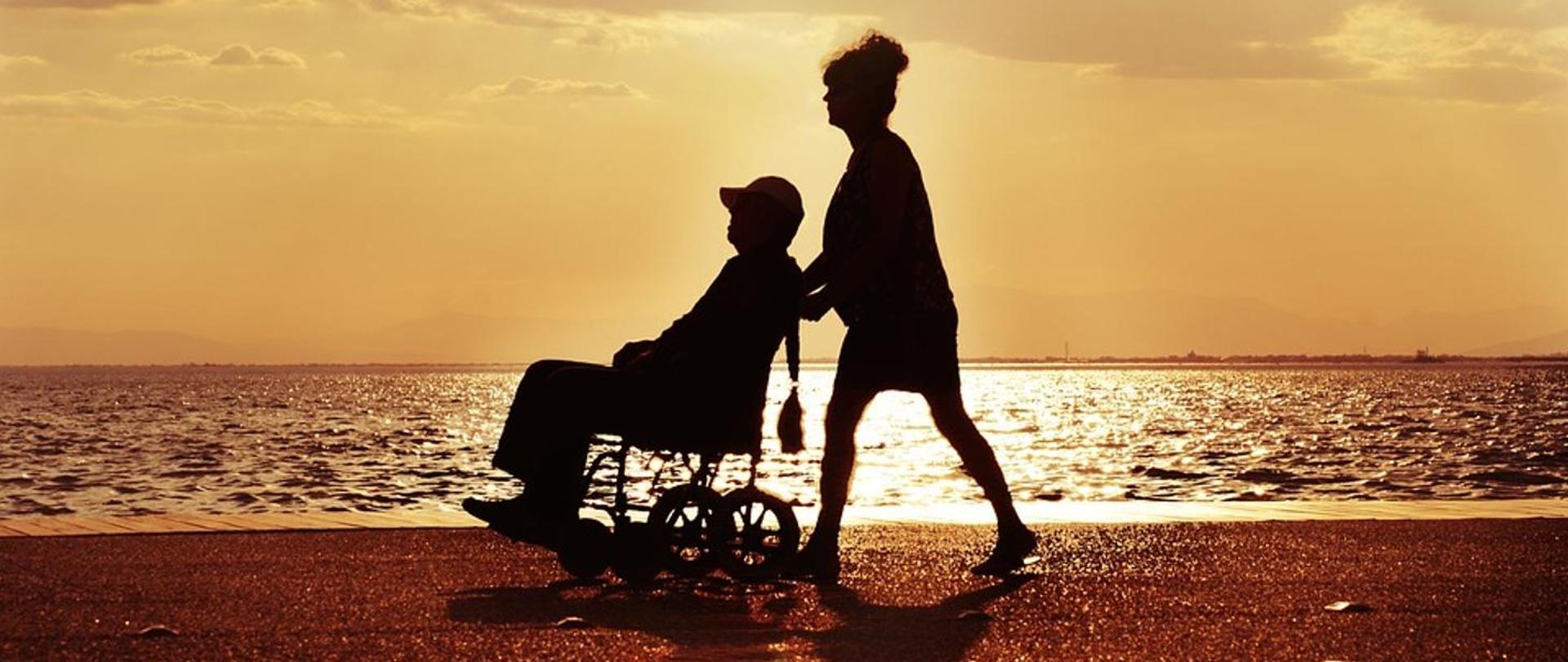 kobieta prowadzi wózek po piasku na którym jest osoba niepełnosprawna w tle morze. Podczas zachodu słońca