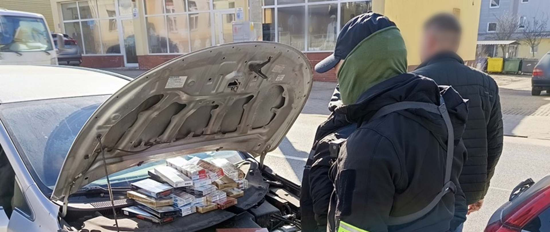 Dwie osoby stoją przed samochodem z otwartą przednią maską. W komorze silnika leżą paczki papierosów. 