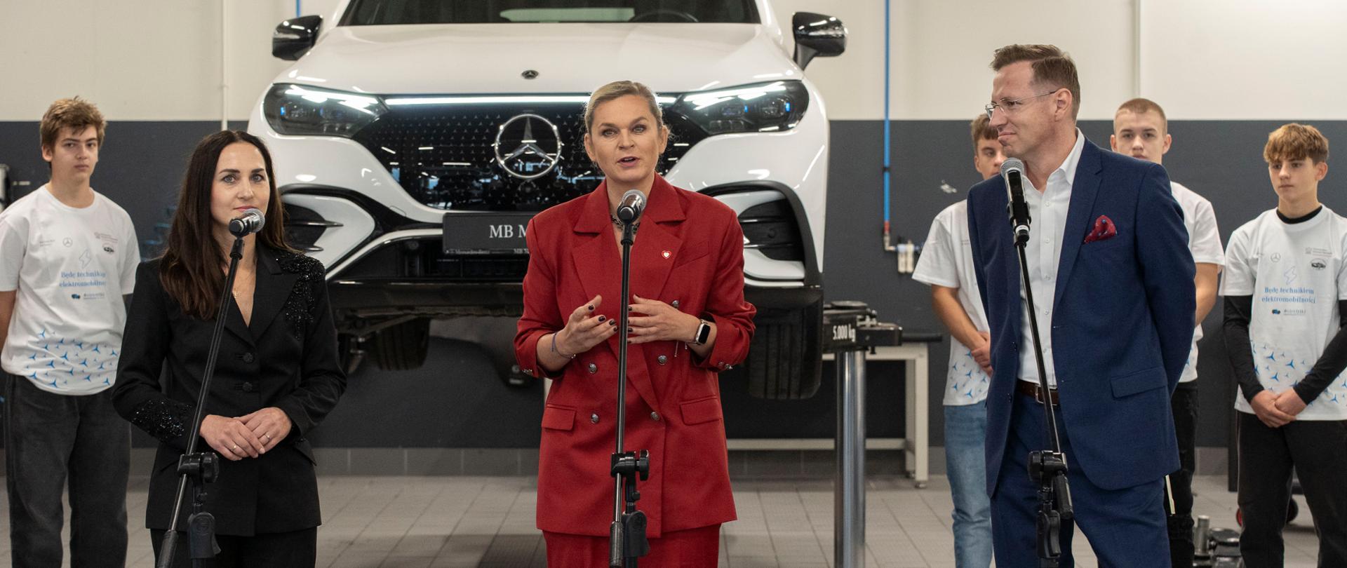 Trzy osoby (dwie kobiety, jeden mężczyzna) stoją przy mikrofonach w warsztacie samochodowym. W tle widoczny jest biały samochód podniesiony na podnośniku. Kobieta w środku w bordowym garniturze gestykuluje.