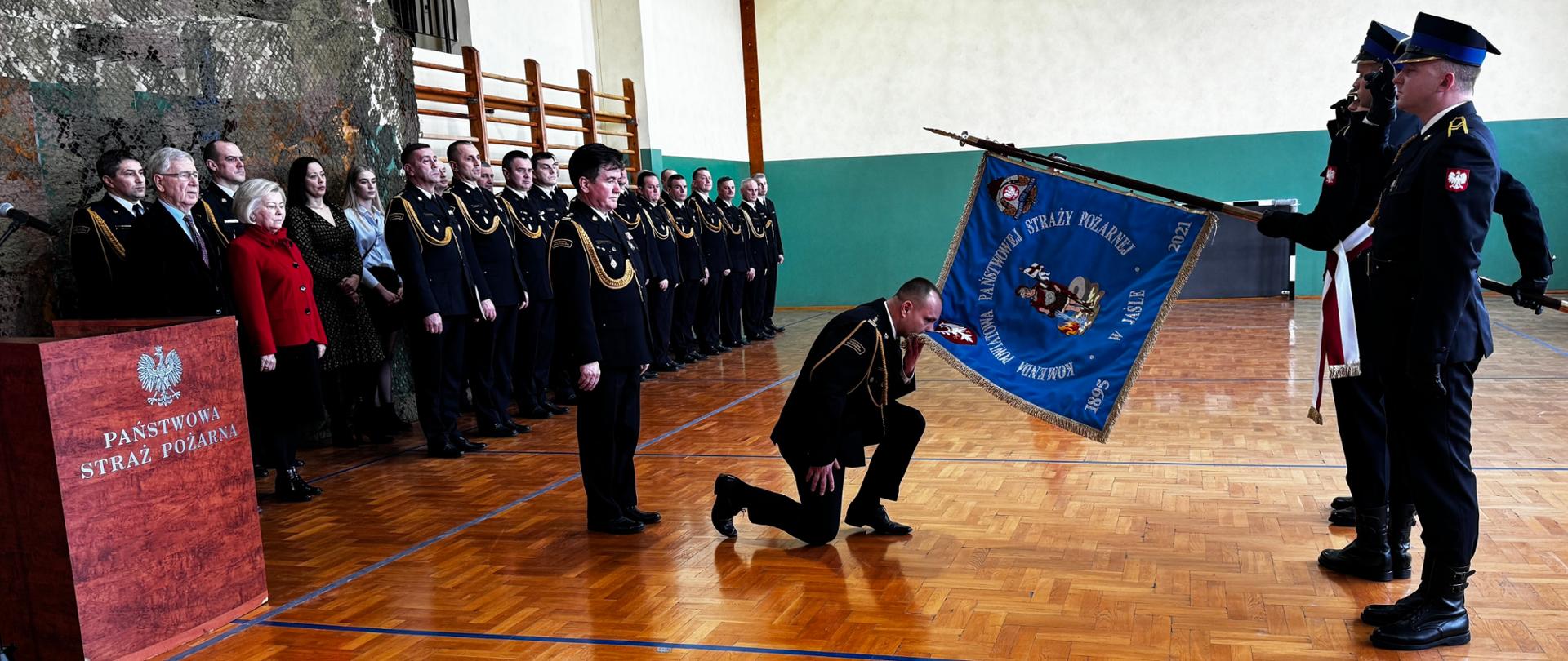 Zdjęcie wewnątrz sali gimnastycznej, w której odbywa się uroczysty apel. Komendant powiatowy żegna się ze sztandarem. W tle stoją zaproszeni goście.