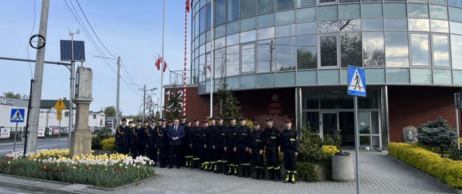 Zdjęcie przedstawia pracowników Komendy Państwowej Straży Pożarnej w Bochni w obecności starosty bocheńskiego Adama Korty. Zdjęcie wykonano pod masztem flagowym z powiewającą flagą narodową. W tle widoczna rotunda budynku PSP w Bochni. 